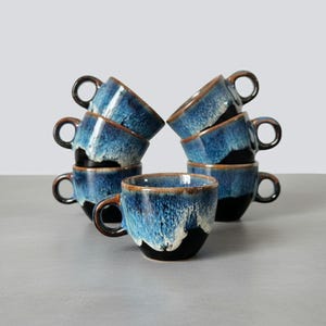 May include: A set of eight ceramic coffee cups. The cups are stacked in two groups, each with a unique design. The cups have a dark blue glaze with white and black accents and brown rims and handles. The cups are on a gray surface.