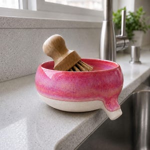 May include: A pink and white ceramic dish with a wooden handled brush. The dish has a spout and is sitting on a countertop next to a stainless steel sink. The brush is made of wood and bristles.