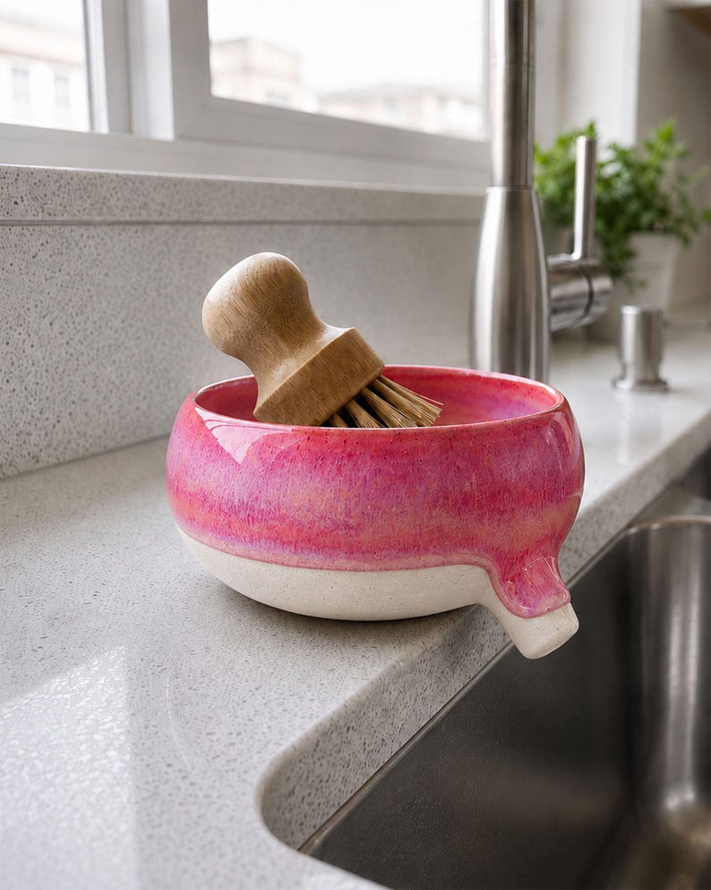 May include: A pink and white ceramic dish with a wooden handled brush. The dish has a spout and is sitting on a countertop next to a stainless steel sink. The brush is made of wood and bristles.