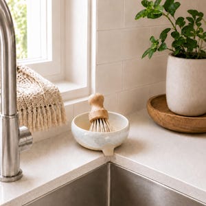 May include: A kitchen sink with a stainless steel faucet and a ceramic dish with a wooden-handled brush. A small potted plant sits on a wooden tray. A window with a cream-colored crocheted cloth is in the background.