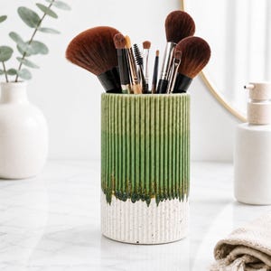 May include: A cylindrical ceramic makeup brush holder with a vertical ribbed texture. The exterior features a two-tone glaze with a vibrant green top section that transitions into a speckled white base, filled with various brown-bristled brushes.