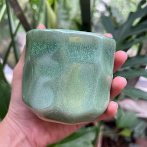 May include: A small, cylindrical ceramic cup with a faceted, geometric exterior surface. The glaze features a variegated finish with shades of sage and mint green, showing a subtle speckled texture and a smooth, glossy sheen across the entire body.