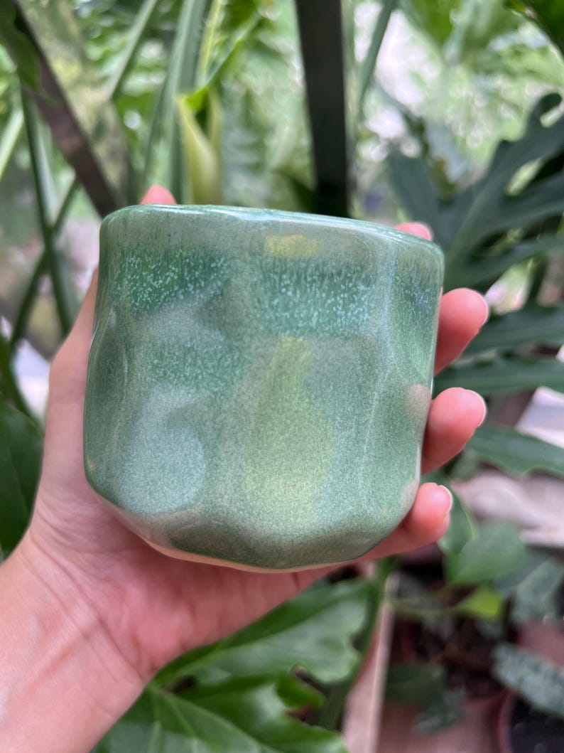 May include: A small, cylindrical ceramic cup with a faceted, geometric exterior surface. The glaze features a variegated finish with shades of sage and mint green, showing a subtle speckled texture and a smooth, glossy sheen across the entire body.