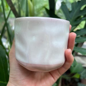 May include: A small, hand-held ceramic cup featuring a glossy white finish on the upper portion and a soft, pale pink glaze on the lower section. The vessel has a distinctively dimpled, uneven surface texture throughout its cylindrical body.