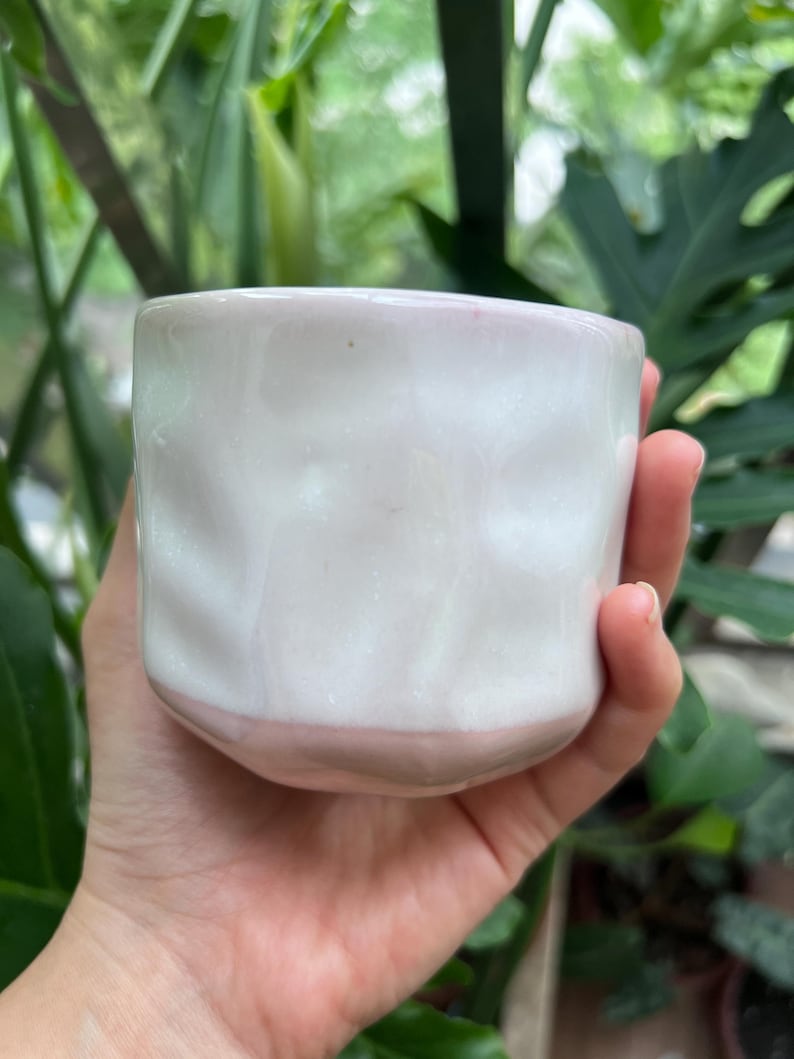 May include: A small, hand-held ceramic cup featuring a glossy white finish on the upper portion and a soft, pale pink glaze on the lower section. The vessel has a distinctively dimpled, uneven surface texture throughout its cylindrical body.