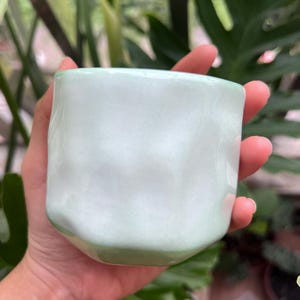 May include: A small, cylindrical ceramic cup with a smooth, glossy finish in a pale mint green color. The exterior surface features subtle, irregular indentations and a wavy, organic texture that creates a soft, rippled appearance around the body.