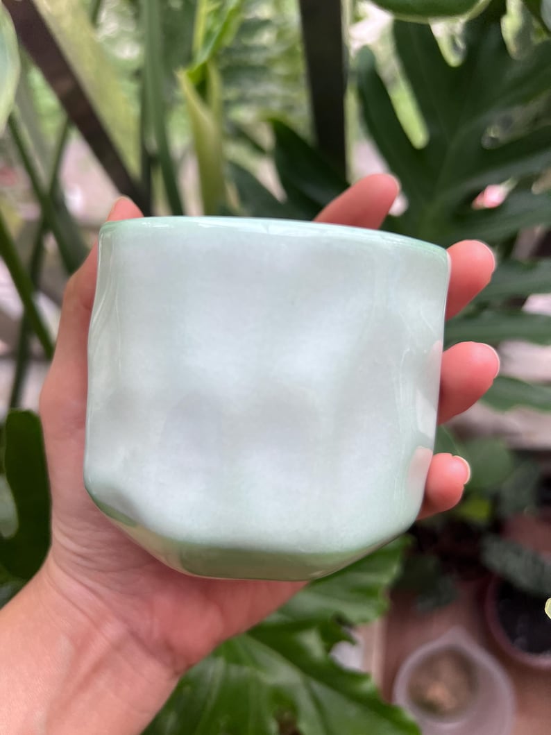 May include: A small, cylindrical ceramic cup with a smooth, glossy finish in a pale mint green color. The exterior surface features subtle, irregular indentations and a wavy, organic texture that creates a soft, rippled appearance around the body.