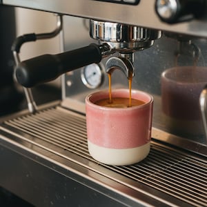 May include: A pink and white ceramic coffee mug is filled with espresso as it brews from a stainless steel espresso machine. The coffee streams into the mug, creating a rich, dark brown color. The mug sits on a metal grate.