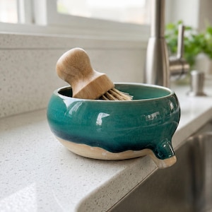 May include: A teal ceramic dish with a spout, holding a wooden-handled brush with natural bristles. The dish has a two-tone design, with a light beige base and a glossy teal glaze. The brush is made of light-colored wood.