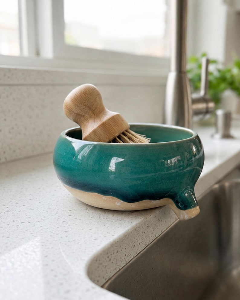 May include: A teal ceramic dish with a spout, holding a wooden-handled brush with natural bristles. The dish has a two-tone design, with a light beige base and a glossy teal glaze. The brush is made of light-colored wood.