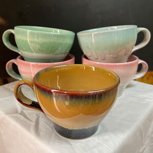 May include: A collection of ceramic coffee mugs in various colors. The mugs are stacked, showcasing a range of hues including green, pink, and a warm, earthy brown with a dark blue base. Each mug has a handle.