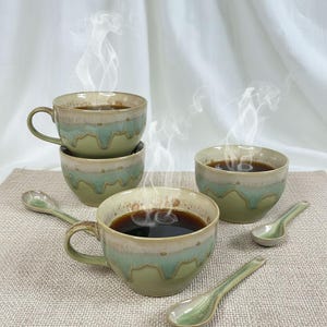 May include: A set of ceramic coffee cups and spoons. The cups are green with a cream-colored rim and a wavy, light green design. The spoons match the cups. Steam rises from the dark coffee in the cups, suggesting a warm beverage.