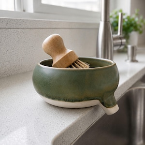 May include: A green ceramic dish with a white rim and a spout, holding a wooden-handled brush with natural bristles. The dish is on a countertop next to a stainless steel sink and faucet.