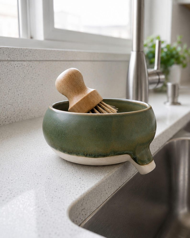 May include: A green ceramic dish with a white rim and a spout, holding a wooden-handled brush with natural bristles. The dish is on a countertop next to a stainless steel sink and faucet.