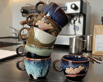 May include: A stack of six ceramic coffee mugs with a dark, glossy finish. The mugs feature a variety of colors, including blue, brown, and green, with a floral design on some. Each mug has a rounded handle.
