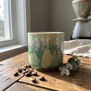 May include: A handmade ceramic mug with a green and white glaze, sitting on a wooden surface. Coffee beans are scattered around the mug. A coffee dripper and a sprig of eucalyptus are in the background.