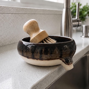 May include: A ceramic kitchen sink brush holder with a wooden brush. The holder is a dark brown with a white base and a spout. The wooden brush has a rounded handle and bristles. The holder is on a white countertop next to a stainless steel sink.