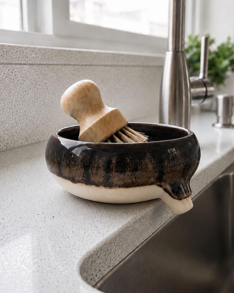 May include: A ceramic kitchen sink brush holder with a wooden brush. The holder is a dark brown with a white base and a spout. The wooden brush has a rounded handle and bristles. The holder is on a white countertop next to a stainless steel sink.