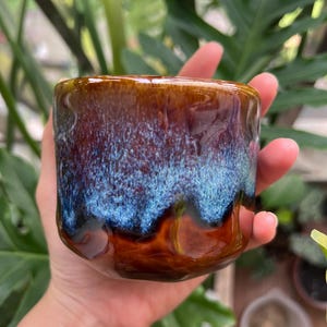 May include: A ceramic mug with a glossy finish featuring a deep brown base color that transitions into a vibrant, textured blue glaze near the rim. The surface has an irregular, undulating shape with visible indentations along the sides.
