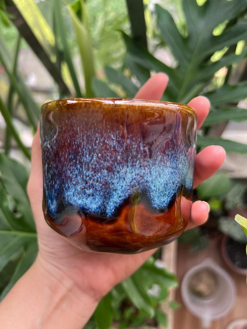 May include: A ceramic mug with a glossy finish featuring a deep brown base color that transitions into a vibrant, textured blue glaze near the rim. The surface has an irregular, undulating shape with visible indentations along the sides.