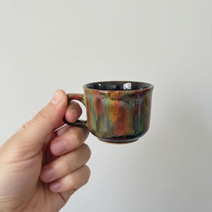 May include: A small, ceramic coffee cup with a handle. The cup features a speckled glaze in shades of brown, green, orange, and red. The interior is black. The cup is held in a hand.
