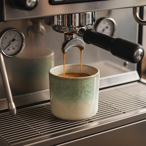 May include: A ceramic mug with a green and white ombre design is filled with freshly brewed espresso. The coffee is pouring from a stainless steel espresso machine. The machine has gauges and a black handle.