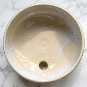 May include: A handmade ceramic bowl with a light beige interior and a glossy, iridescent finish. The bowl has a circular shape with a small drainage hole at the bottom. The rim is a soft, muted green. The bowl is likely for a bathroom sink.