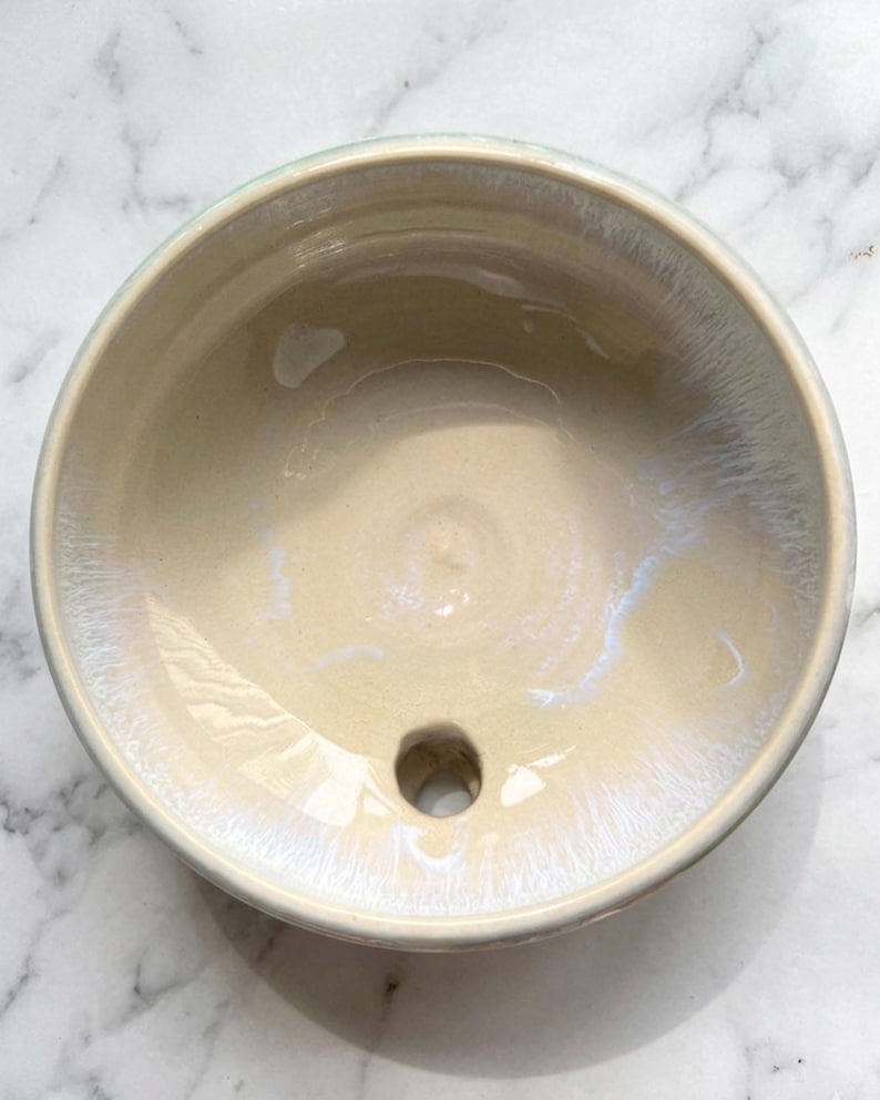 May include: A handmade ceramic bowl with a light beige interior and a glossy, iridescent finish. The bowl has a circular shape with a small drainage hole at the bottom. The rim is a soft, muted green. The bowl is likely for a bathroom sink.
