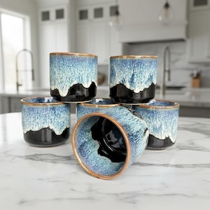 May include: A set of six ceramic tumblers with a unique glaze. The cups feature a gradient of colors, including blue, white, and black, with a bronze-colored rim. The design gives the appearance of ocean waves. These tumblers are suitable for beverages.