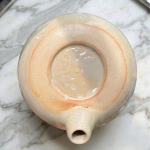 May include: A handmade ceramic teapot with a spout and a circular base. The teapot is primarily cream-colored with a peach-colored ring around the base. The bottom of the teapot is visible, showing the unglazed ceramic.