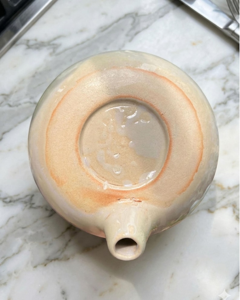 May include: A handmade ceramic teapot with a spout and a circular base. The teapot is primarily cream-colored with a peach-colored ring around the base. The bottom of the teapot is visible, showing the unglazed ceramic.