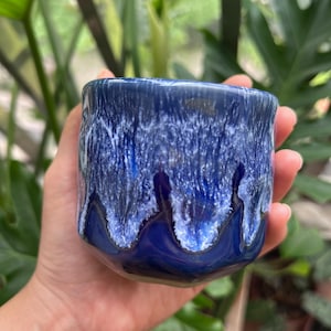 May include: A small ceramic cup featuring a glossy finish with a deep royal blue base color. The upper portion of the cup displays a textured, dripped glaze effect in shades of white and light blue that cascades down over the dark blue surface.