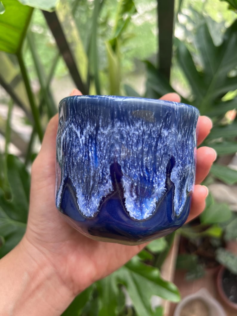 May include: A small ceramic cup featuring a glossy finish with a deep royal blue base color. The upper portion of the cup displays a textured, dripped glaze effect in shades of white and light blue that cascades down over the dark blue surface.