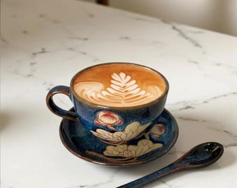 May include: A ceramic coffee cup and saucer set with a floral design. The cup is filled with coffee and has a leaf design in the foam. A matching spoon rests on the table. The set is blue with brown accents.