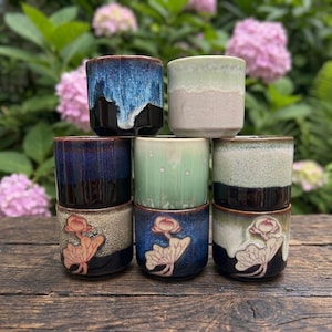 May include: A stack of eight ceramic teacups in various colors, including blue, green, and beige. Some cups feature a floral design. The cups are arranged on a wooden surface with a backdrop of pink flowers.