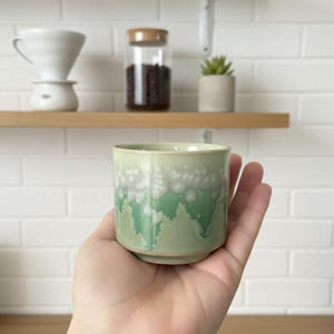May include: A small, handmade ceramic cup with a light green glaze and white, bubble-like accents. The cup is held in a hand, showcasing its unique texture and design. The cup is a perfect size for espresso or a small beverage.
