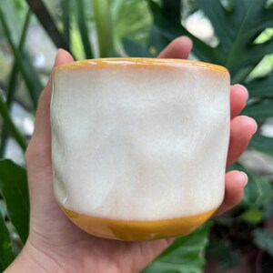 May include: A ceramic mug with a wavy, textured surface featuring a glossy white glaze on the body, contrasted by a solid mustard yellow band at the base and a matching mustard yellow rim. The mug has a rounded, handleless, cylindrical shape.