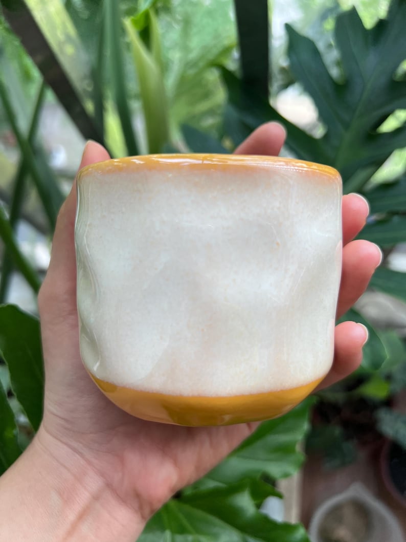 May include: A ceramic mug with a wavy, textured surface featuring a glossy white glaze on the body, contrasted by a solid mustard yellow band at the base and a matching mustard yellow rim. The mug has a rounded, handleless, cylindrical shape.