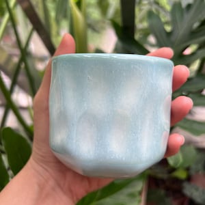 May include: A small, cylindrical ceramic cup with a glossy, light blue glaze. The surface features a subtle, undulating texture with vertical indentations that create a soft, rippled effect around the exterior of the vessel.