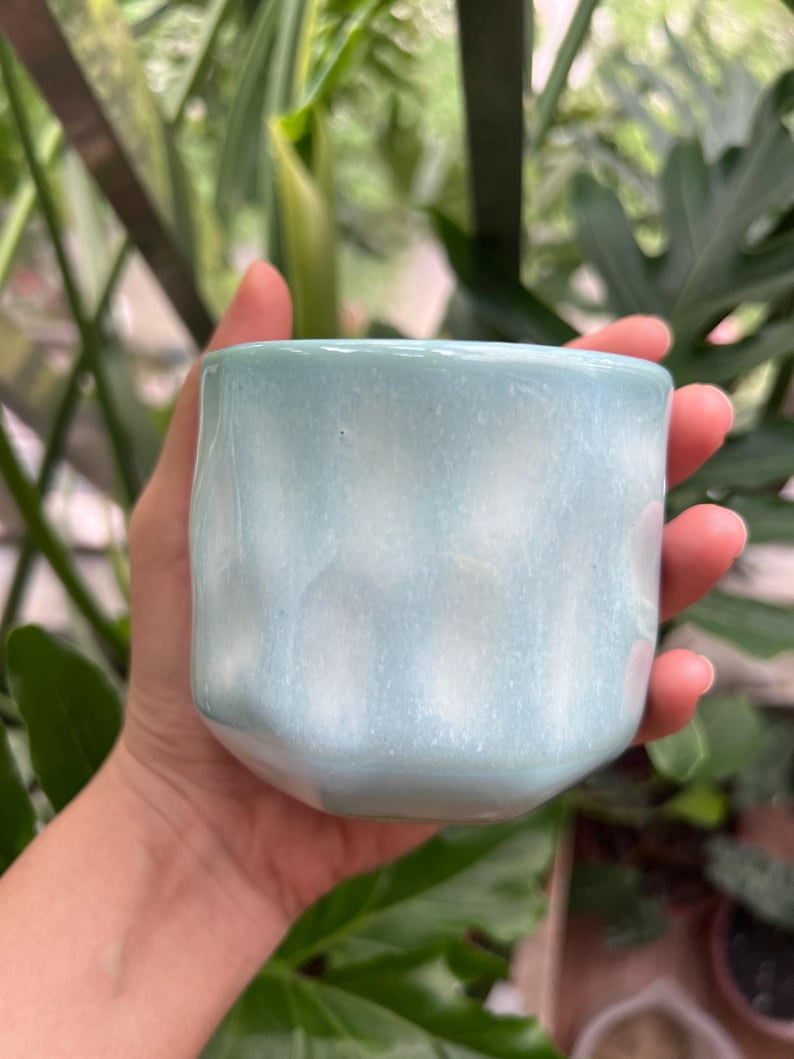May include: A small, cylindrical ceramic cup with a glossy, light blue glaze. The surface features a subtle, undulating texture with vertical indentations that create a soft, rippled effect around the exterior of the vessel.
