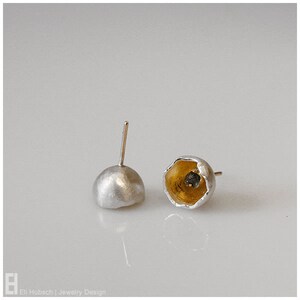 May include: A pair of silver earrings with a textured finish. One earring has a gold-toned center with a small black dot. The earrings are designed in a simple, modern style.
