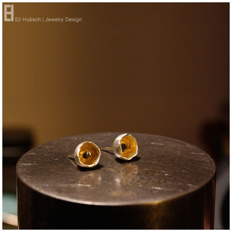 May include: A pair of silver earrings with gold accents. The earrings are shaped like small bowls with a black dot in the center.