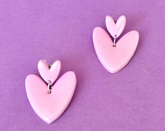 May include: A pair of pink heart-shaped earrings with a smaller heart charm hanging from the larger heart. The earrings are made of polymer clay and have a glossy finish.