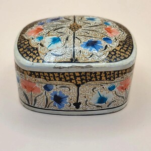 May include: A small, oval-shaped decorative box featuring a hand-painted floral pattern in shades of blue, orange, and white. The surface is adorned with intricate black and gold filigree details, creating a contrast against the light base.
