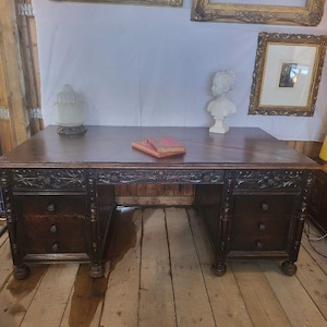Op de afbeelding: Een donkerbruin houten bureau met een rechthoekig blad, twee sokkels met elk drie laden en gedetailleerd houtsnijwerk langs de voorzijde. Op het bureaublad liggen twee gestapelde boeken en een wit borstbeeld aan de rechterkant.