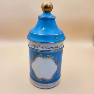 Pode incluir: Um pote cilíndrico de porcelana com tampa apresentando um remate esférico dourado. O pote é pintado em azul brilhante com painéis brancos, decorado com filigranas douradas, motivos de folhas e pequenos detalhes de estrelas na tampa.