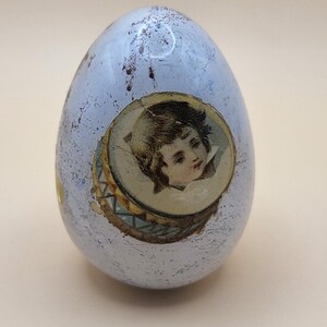 May include: A light blue, egg-shaped decorative object with a distressed finish. It features a circular image of a cherub. The egg is likely a decorative item for Easter or home decor.