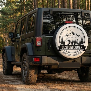 May include: A dark green Jeep SUV parked on a dirt path in a forest. The spare tire cover features a circular design with a bear, mountains, and the text "CUSTOM COVER" and "YOUR LOGO - YOUR TEXT".