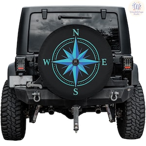 May include: Black spare tire cover featuring a blue and teal compass rose design. The compass rose has the letters N, E, S, and W. The cover is on the back of a black Jeep.