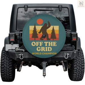Puede incluir: Una funda para rueda de repuesto de color verde azulado con un diseño retro que presenta una silueta de Bigfoot, dos pinos y un sol poniente. El texto "OFF THE GRID WORLD CHAMPION" está impreso en amarillo.